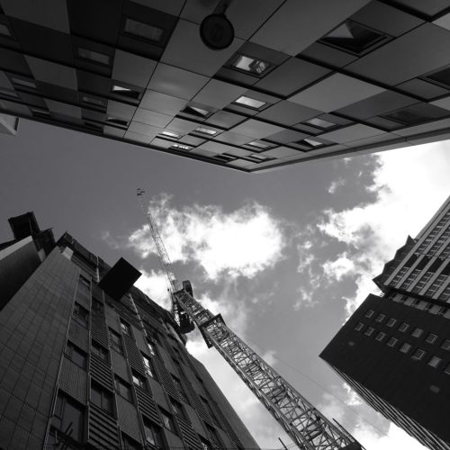 Low angle view of modern skyscrapers and construction crane against cloudy sky in black and white.